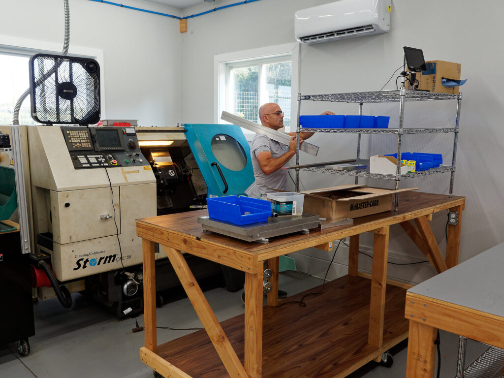 Working man standing at organizational bins on a wire shelf with a small assortment of aluminum and 1045 carbon steel in the foreground. A turret CNC lathe is shown operating in the background, surrounded by clean organized multipurpose tables.