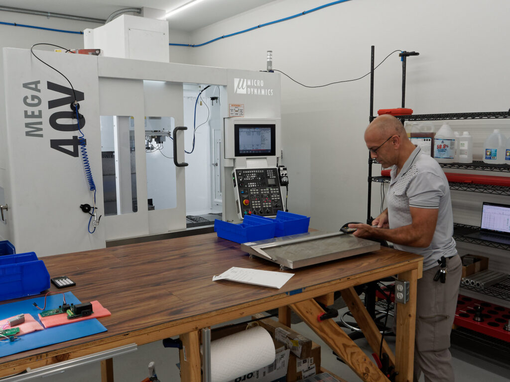 A portion of the machine shop & laboratory showing a large plate of wrought aluminum being assessed with a 1 meter digital caliper, the left corner, and a Microdynamics Mega40V vertical machining center in the background."