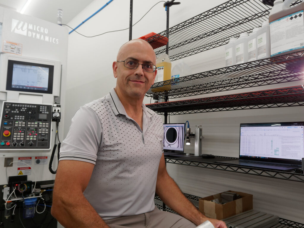 Shop owner located between a Dino-Lite microscope inspection station and the Mitsubishi 830Mv control of a modern VMC, with chemicals on a shelf behind him.